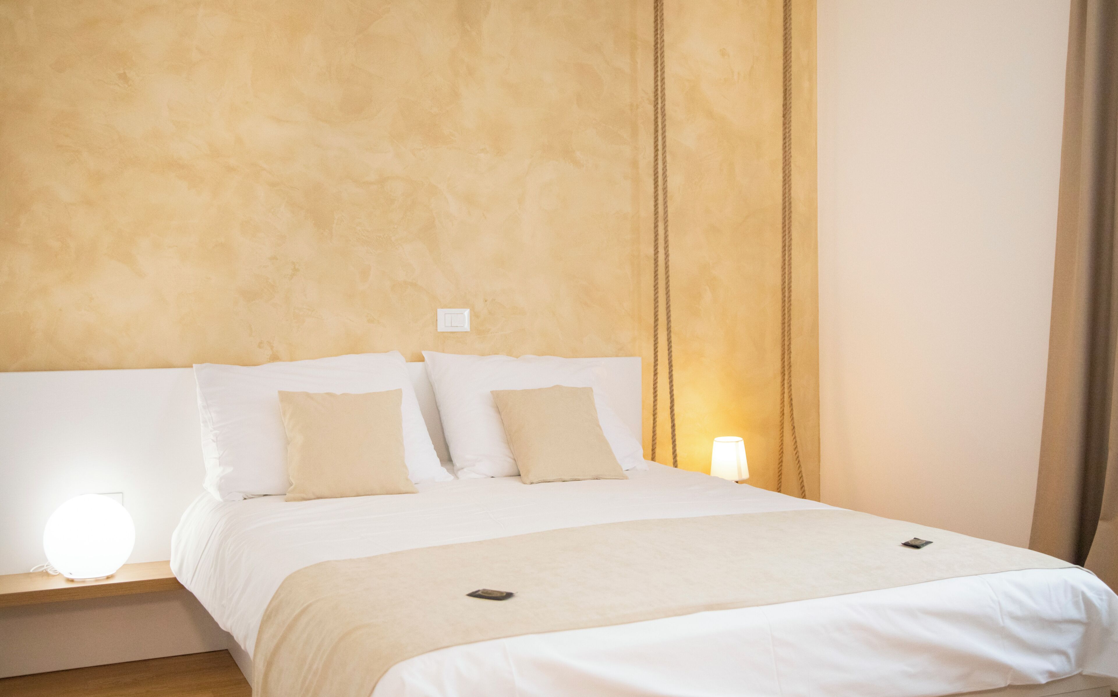 A neatly made bed with white bedding and two pillows, a bedside table with a lamp, and a wall switch.