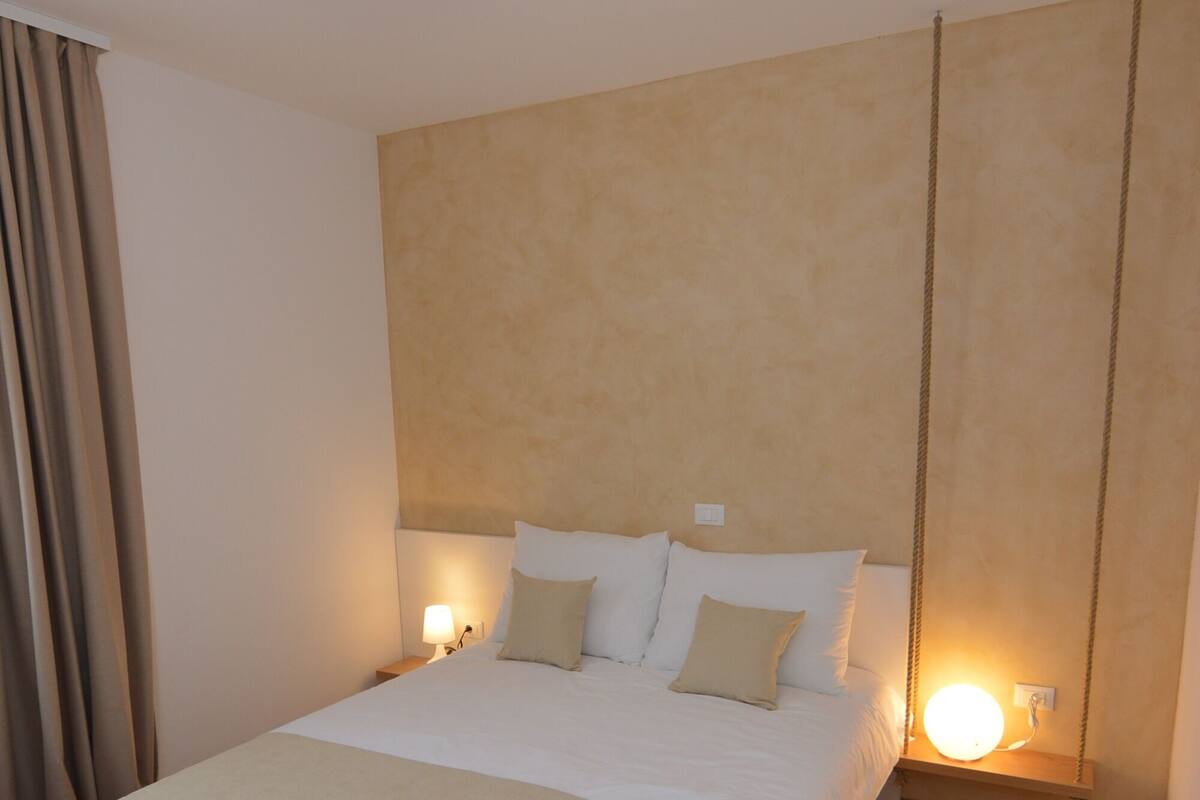 A neatly made bed with white bedding and a beige throw blanket, a bedside lamp, and a hanging swing chair.