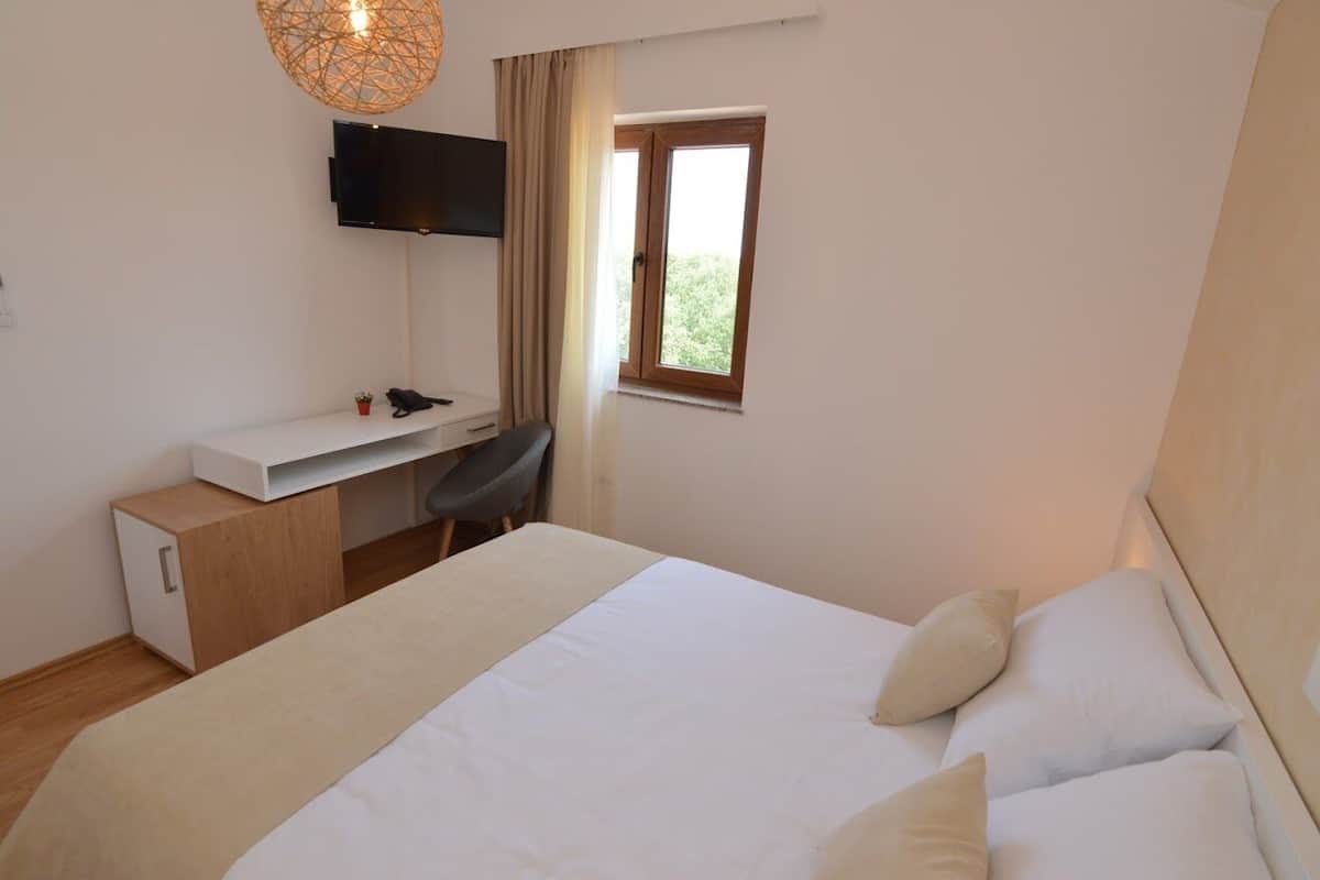 A bedroom with a bed, a desk, a chair, a TV, and a window.