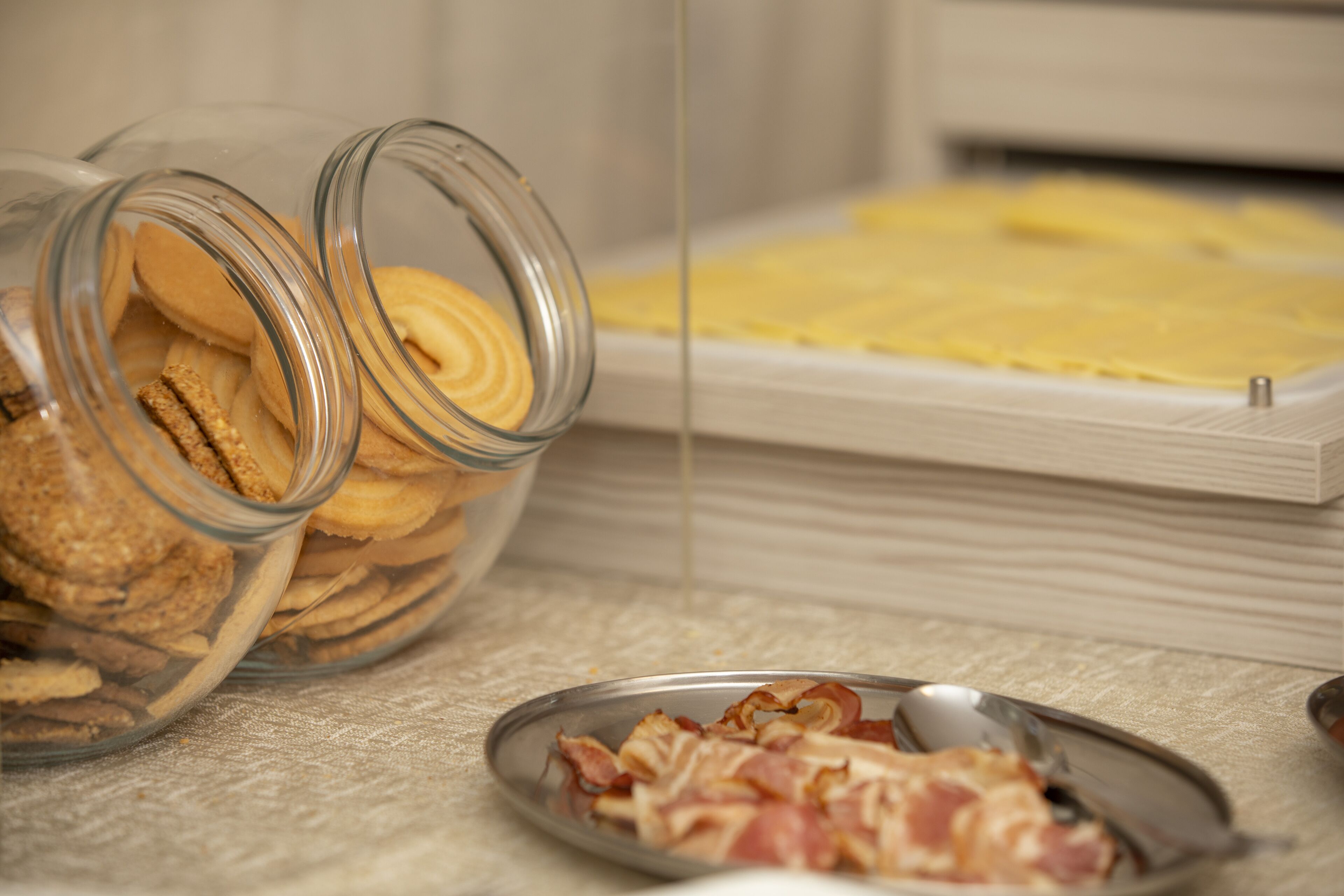 Een ontbijttafel met een pot koekjes, een bord spek en een houten snijplank met een gele spread.
