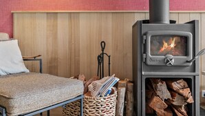 A room with a wood-burning stove, a chair, a table, and a basket of firewood. - Blue Haven at Aireys (Aireys Inlet)