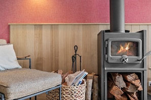 A room with a wood-burning stove, a chair, a table, and a basket of firewood. - Blue Haven at Aireys (Aireys Inlet)