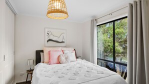 A bedroom with a bed, a bedside table, a lamp, a hanging light fixture, a window with curtains, and a wall-mounted artwork. - Blue Haven at Aireys (Aireys Inlet)
