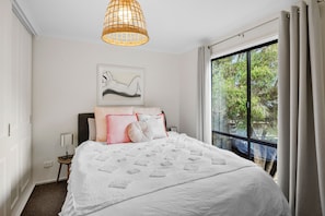 A bedroom with a bed, a bedside table, a lamp, a hanging light fixture, a window with curtains, and a wall-mounted artwork. - Blue Haven at Aireys (Aireys Inlet)