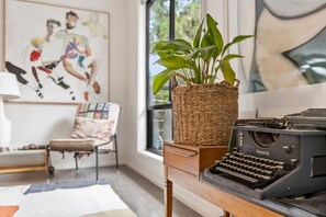 A room with a vintage typewriter on a wooden desk, a wicker basket with a plant, a colorful armchair, and a painting on the wall. - Blue Haven at Aireys (Aireys Inlet)