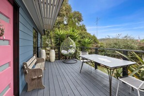A wooden deck with a table and chairs, surrounded by greenery and a pink building. - Blue Haven at Aireys (Aireys Inlet)