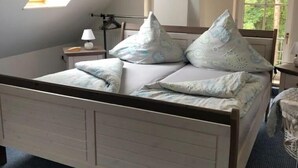 Ein Schlafzimmer mit schrägen Wänden, einem Dachfenster, einem Bett mit gemusterten Kissen, einem Nachttisch mit Lampe und einem Fenster mit Blick ins Grüne.