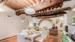 A dining area with a white table set for four, surrounded by white chairs. A chandelier hangs from the ceiling, and a brick archway is visible in the background. - Casa Al Dipinto in Lucca (Lucca)