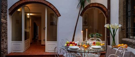 An outdoor dining area with a glass table and white metal chairs, surrounded by brick walls and arched doorways.