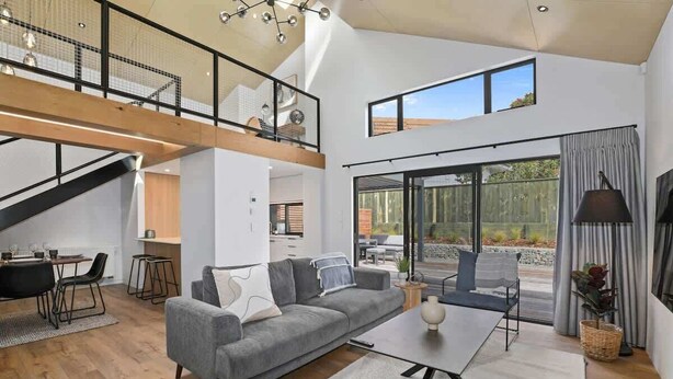 A spacious living area with a grey sofa, a coffee table, and a dining area with a table and chairs. There is a staircase leading to an upper level, and large windows providing natural light. - Serenity on Sophia - Spa Pool, Sleeps 6 (Rotorua)