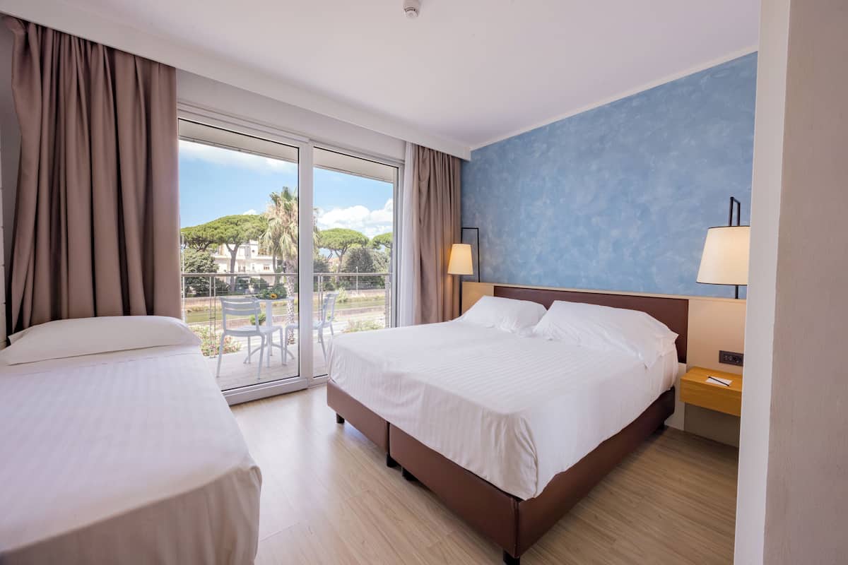 Une chambre d’hôtel avec un grand lit, un lit plus petit, un balcon avec vue et un bureau avec une lampe.