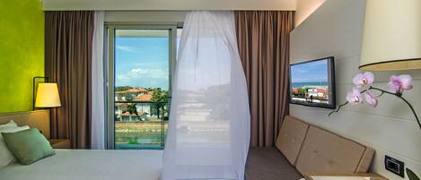 A hotel room with a bed, a desk, a chair, a TV, and a view of the beach.