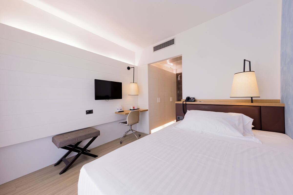 Une chambre d’hôtel moderne équipée d’un grand lit, d’un bureau, d’une chaise, d’une télévision et de deux lampes fixées au mur.