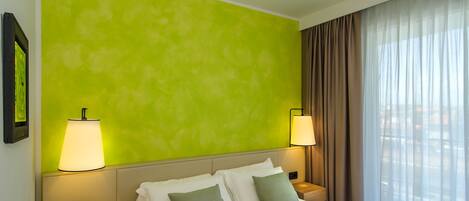 Una habitación de hotel con una pared verde brillante, una cama con ropa blanca y almohadas verdes, dos lámparas de noche, una mesita de noche con un teléfono y una ventana con cortinas translúcidas.