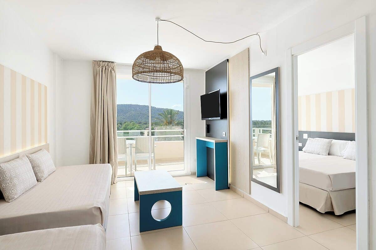 A modern hotel room with a bed, a sofa, a TV, and a balcony with a view of the hills.