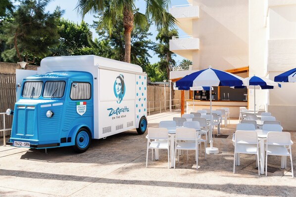 Un camión de comida azul con la marca 'Zia Rosetta On the Road' estacionado frente a un edificio con mesas al aire libre y sombrillas.