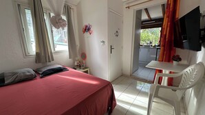 A bedroom with a bed, a desk, and a chair. There is a window with curtains, a door, and a view of an outdoor area. - Appart Bellissima Calme Paisible Entouré de Verdure (Baie-Mahault)