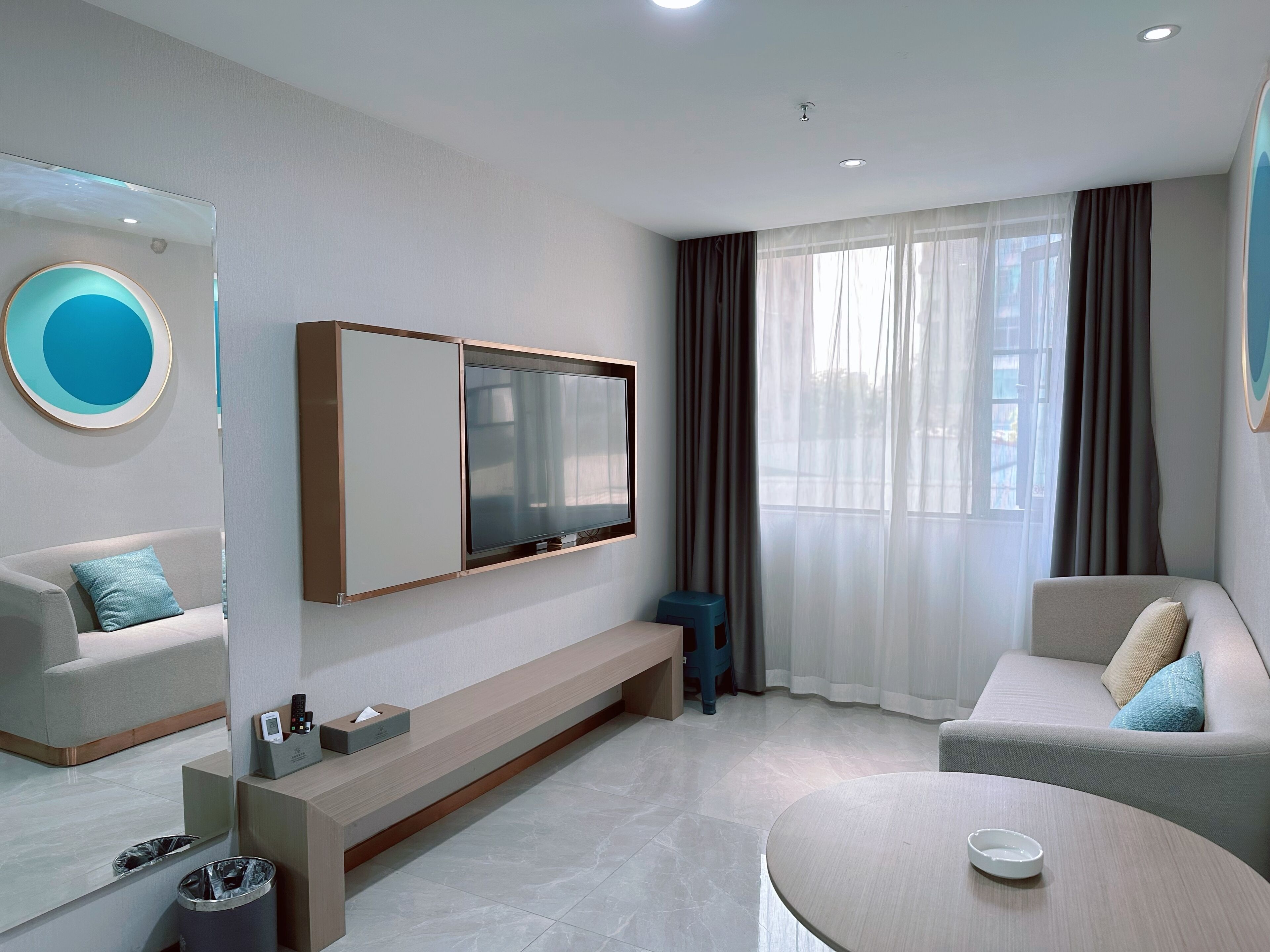 A modern hotel room with a large flat-screen TV mounted on the wall, a sofa, a small round table, and a trash bin.