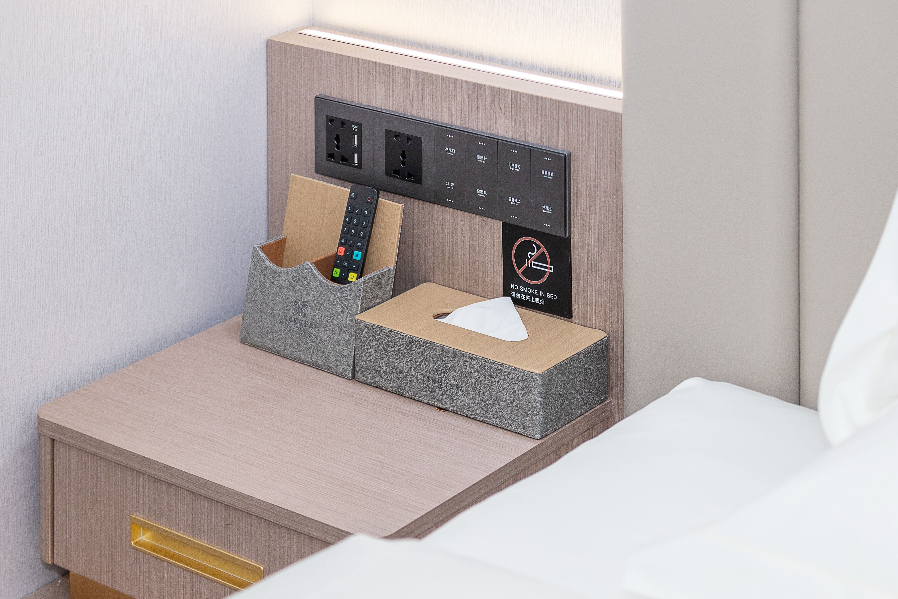 Une table de chevet bien rangée, avec un support pour télécommande, une boîte de mouchoirs et un panneau «interdit de fumer».