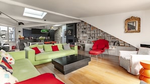 Un vaste espace de vie agrémenté d’un canapé vert, d’un fauteuil rouge et d’une table basse noire. Un escalier mène à une bibliothèque remplie de livres.