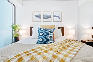 Habitación de hotel con cama, un ventanal grande, una planta en maceta y tres cuadros con imágenes de ballenas en la pared.