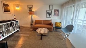 Una sala de estar con un sofá marrón, una mesita redonda pequeña, una estantería con CDs, una lámpara de pie y obras de arte enmarcadas en las paredes.