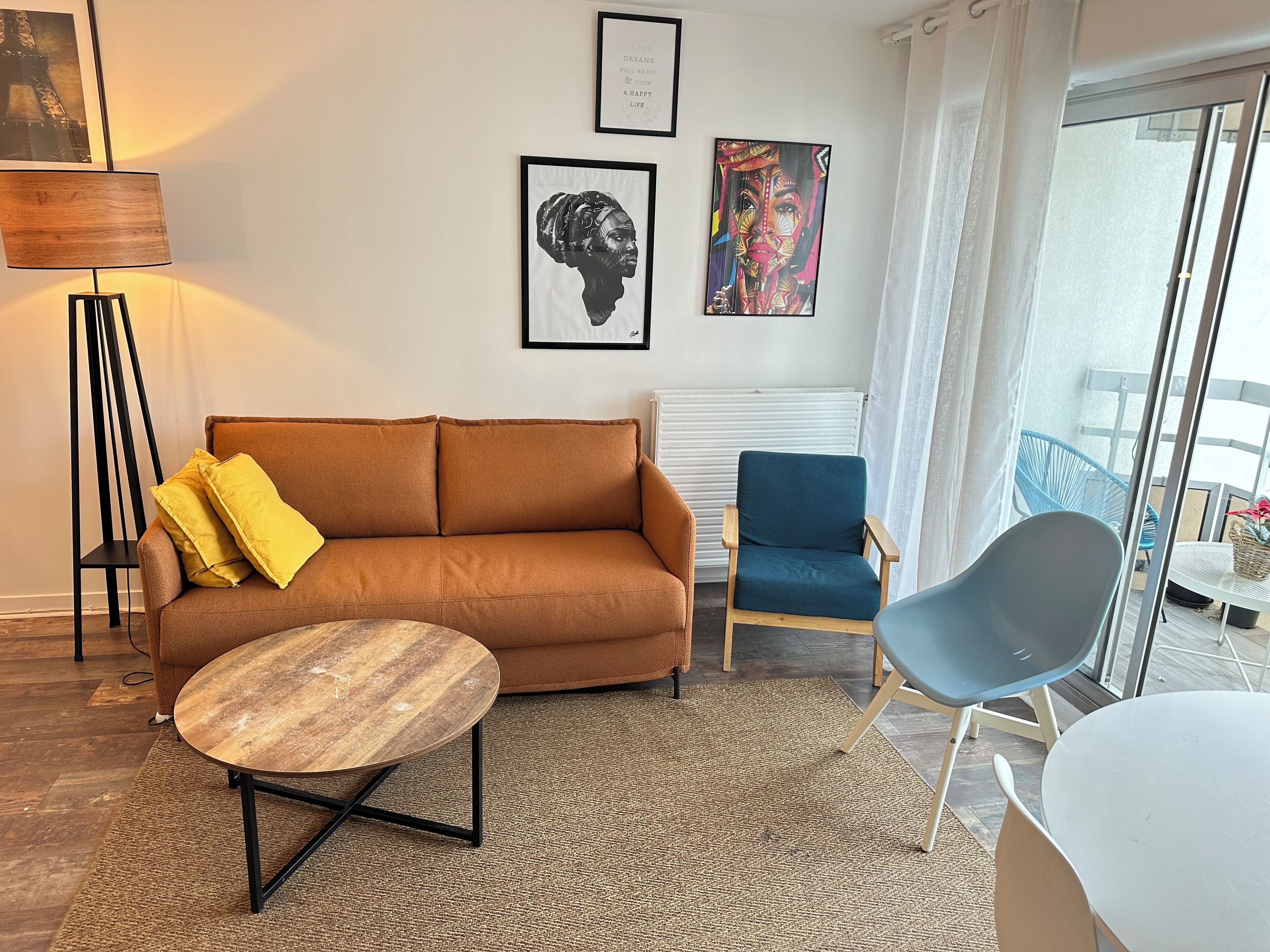 A modern living room with a brown sofa, a round wooden coffee table, a blue armchair, and a white chair. There are framed artworks on the wall and a floor lamp.