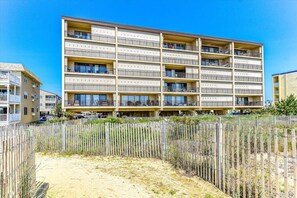 A multi-story hotel with balconies, a sandy ground in front, and a wooden fence.