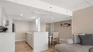 A modern kitchen with white cabinetry, a dining area with a table and chairs, and a grey sofa in a neutral-toned living space. - Completely Renovated Three Bedroom Oceanfront Condo (Ocean City)