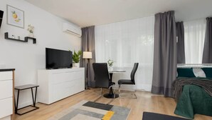A modern hotel room with a flat-screen TV, a desk with a chair, a bed with a green blanket, and a large window with grey curtains. - Air-conditioned Studio with a Balcony | Close to Pola Mokotowskie (Warszawa)