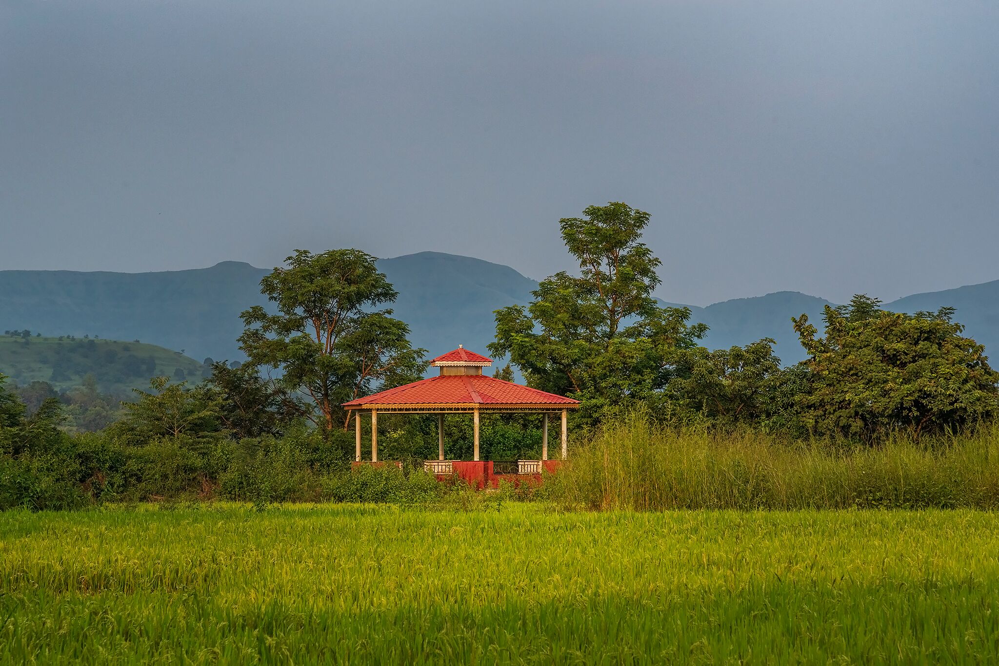 Um gazebo com telhado vermelho em um campo gramado, cercado por árvores e colinas.