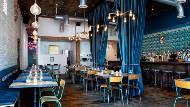 A restaurant with long communal tables, blue curtains, and a bar area with stools.