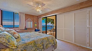 A bedroom with a bed, a ceiling fan, a large window with blinds, and a view of the ocean.