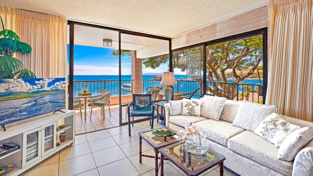 A spacious living room with a large sofa, a coffee table, and a TV on a stand. There is a balcony with a view of the ocean and a painting of a beach scene.