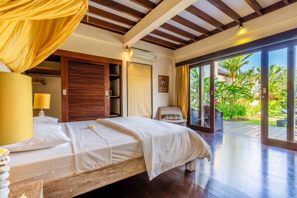Une chambre spacieuse avec un grand lit, un plafond en bois et une vue sur un jardin luxuriant, accessible par une porte-fenêtre coulissante.