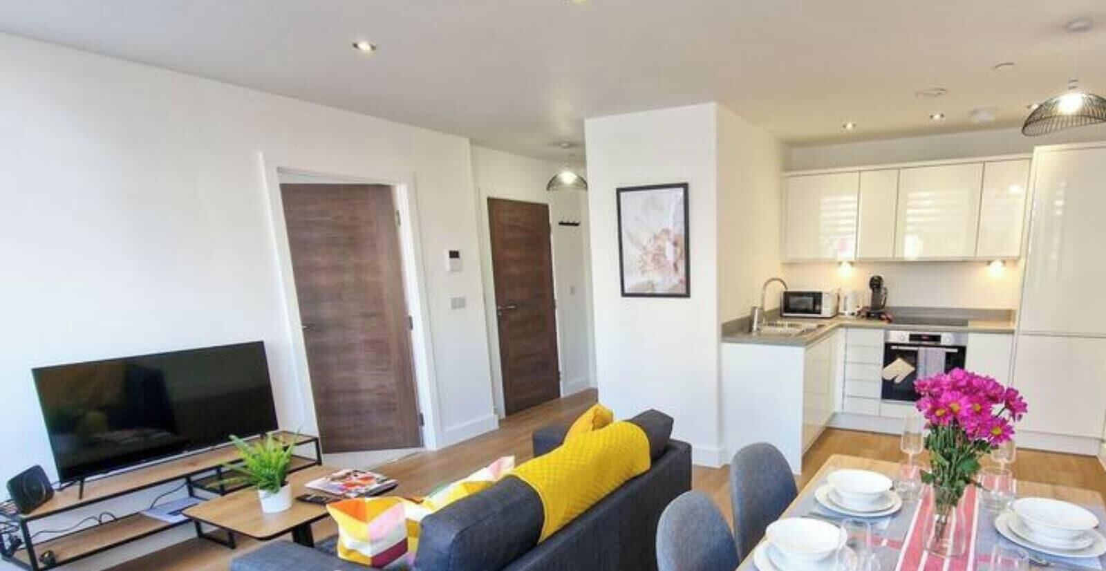 A modern living room with a flat-screen TV, a sofa with a yellow pillow, a dining table set for two, and a kitchen with white cabinets and a microwave.