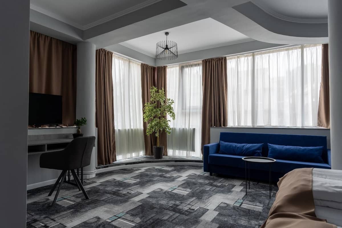 Een moderne hotelkamer met een blauwe bank, een bureau met een televisie, een potplant en grote ramen met gordijnen.