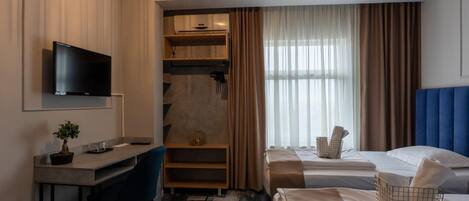 A hotel room with a desk, chair, two beds, a TV, a small plant, and a window with curtains.