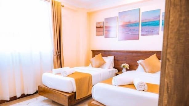 A hotel room with two beds, a wooden headboard, a white rug, and a view of the beach through the window. - Kije Place Hotel (Nairobi)