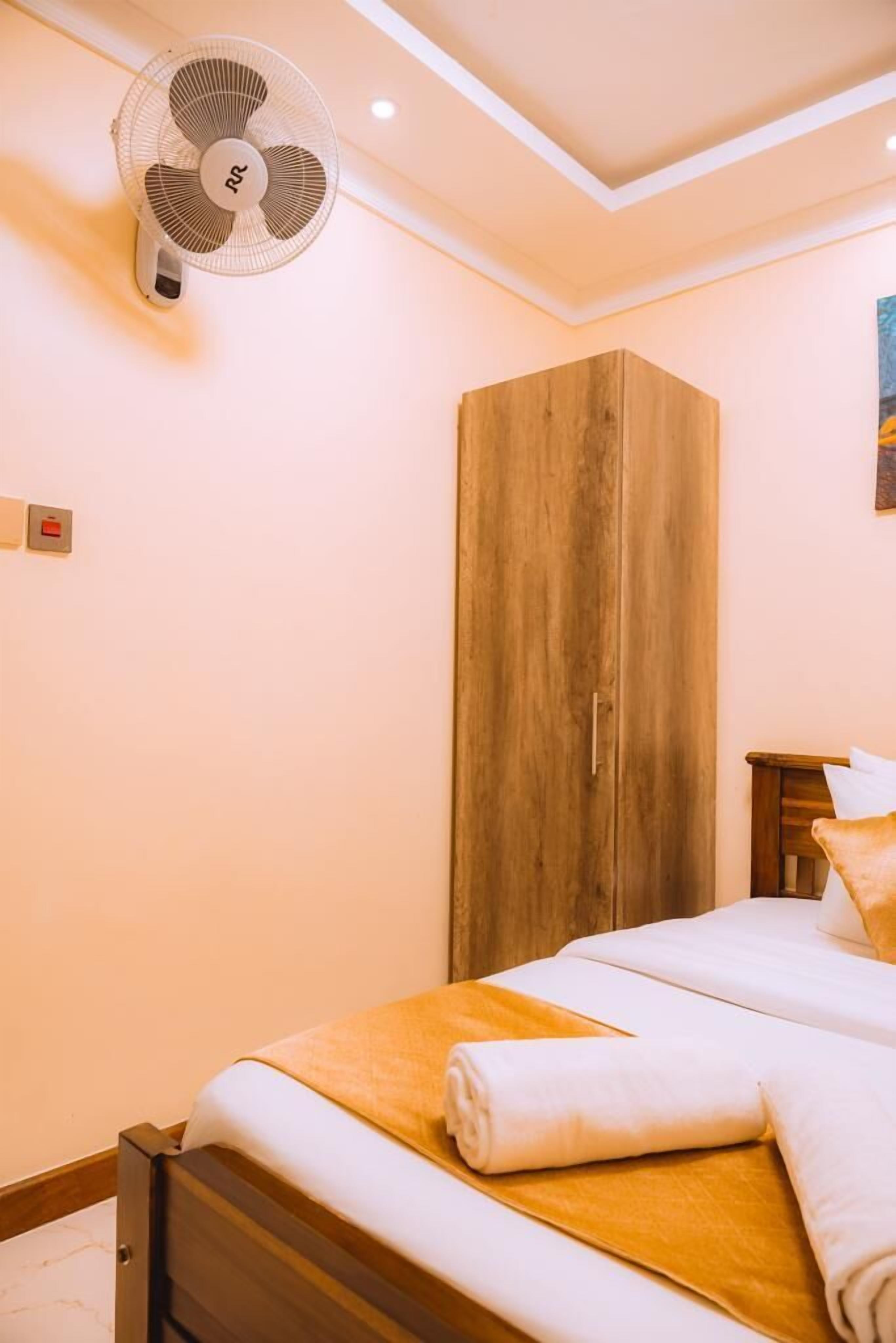 Een hotelkamer met een bed, een houten garderobe, een nachtkastje en een plafondventilator.