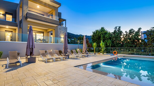 A modern poolside area with lounge chairs and umbrellas, a clear pool, and a building with balconies. - Chris & Stratos villa with private pool and playground for kids! (Chania)