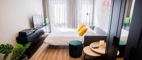 A hotel room with a bed, a desk, a chair, a small table, and a potted plant.