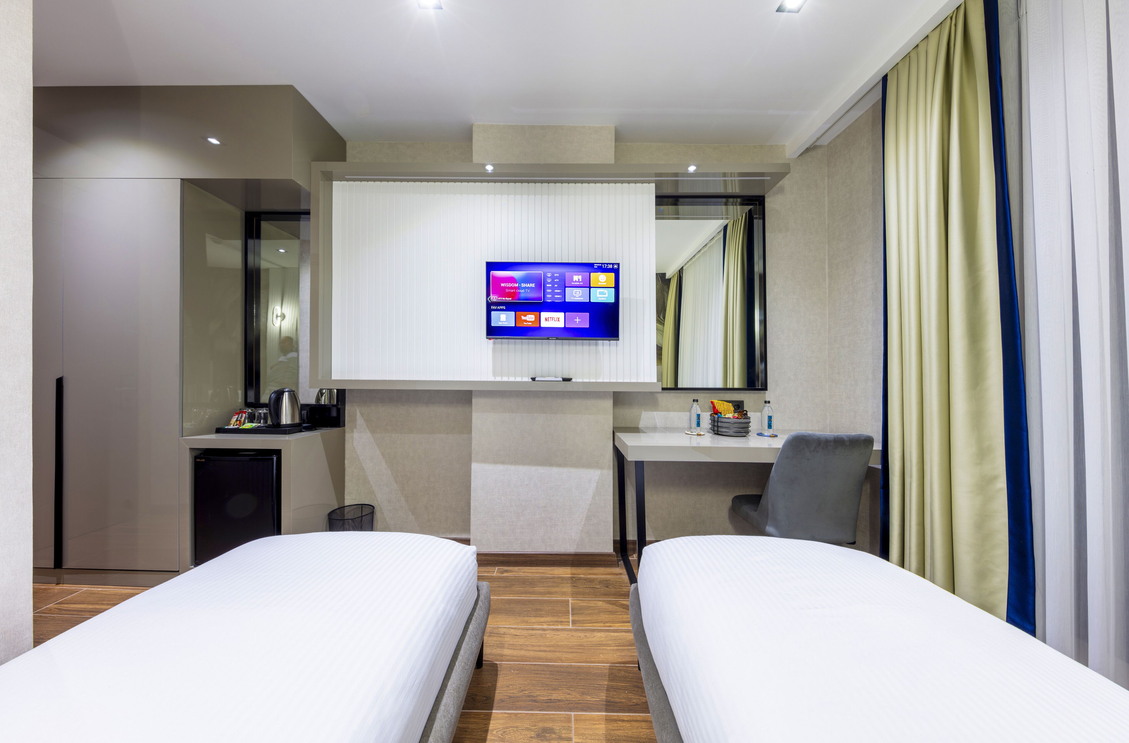 A hotel room with two beds, a desk with a chair, a small kitchenette, and a TV mounted on the wall.