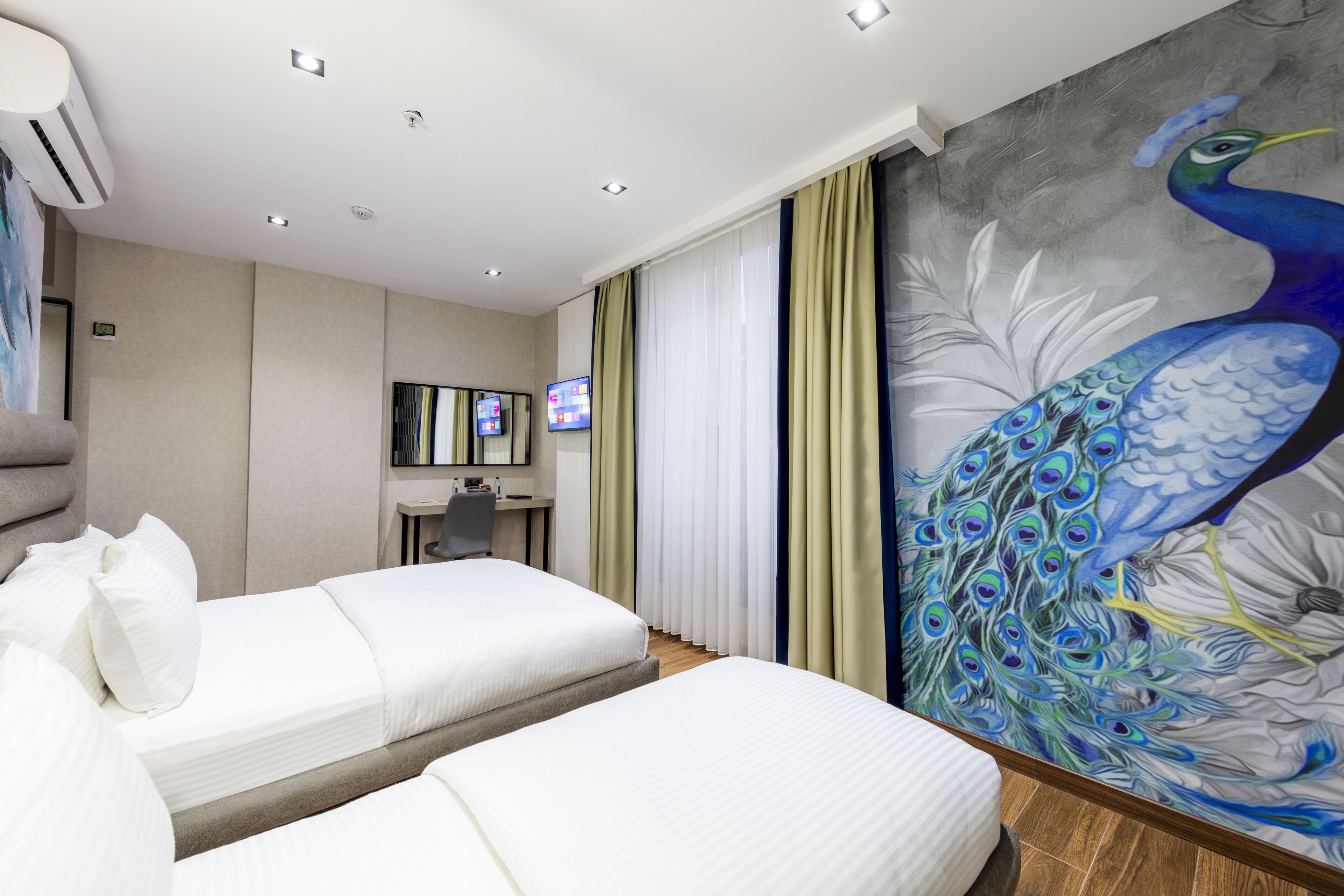 Habitación de hotel con dos camas, un escritorio, un televisor y un mural con un pavo real.