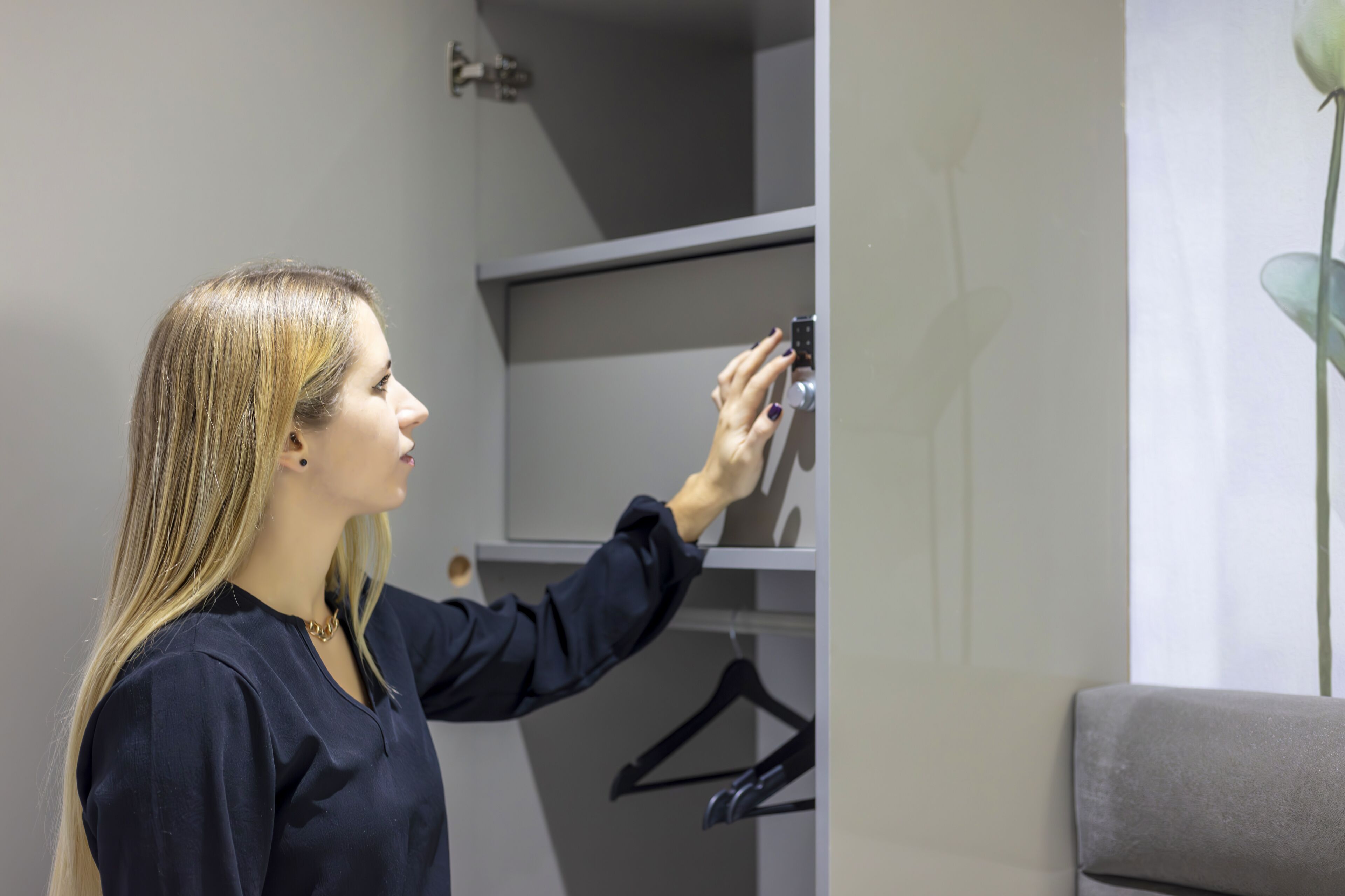 Une personne prend une photo avec un téléphone intelligent dans une pièce dont le mur est gris, où se trouve une chaise grise et un cintre noir accroché au mur.