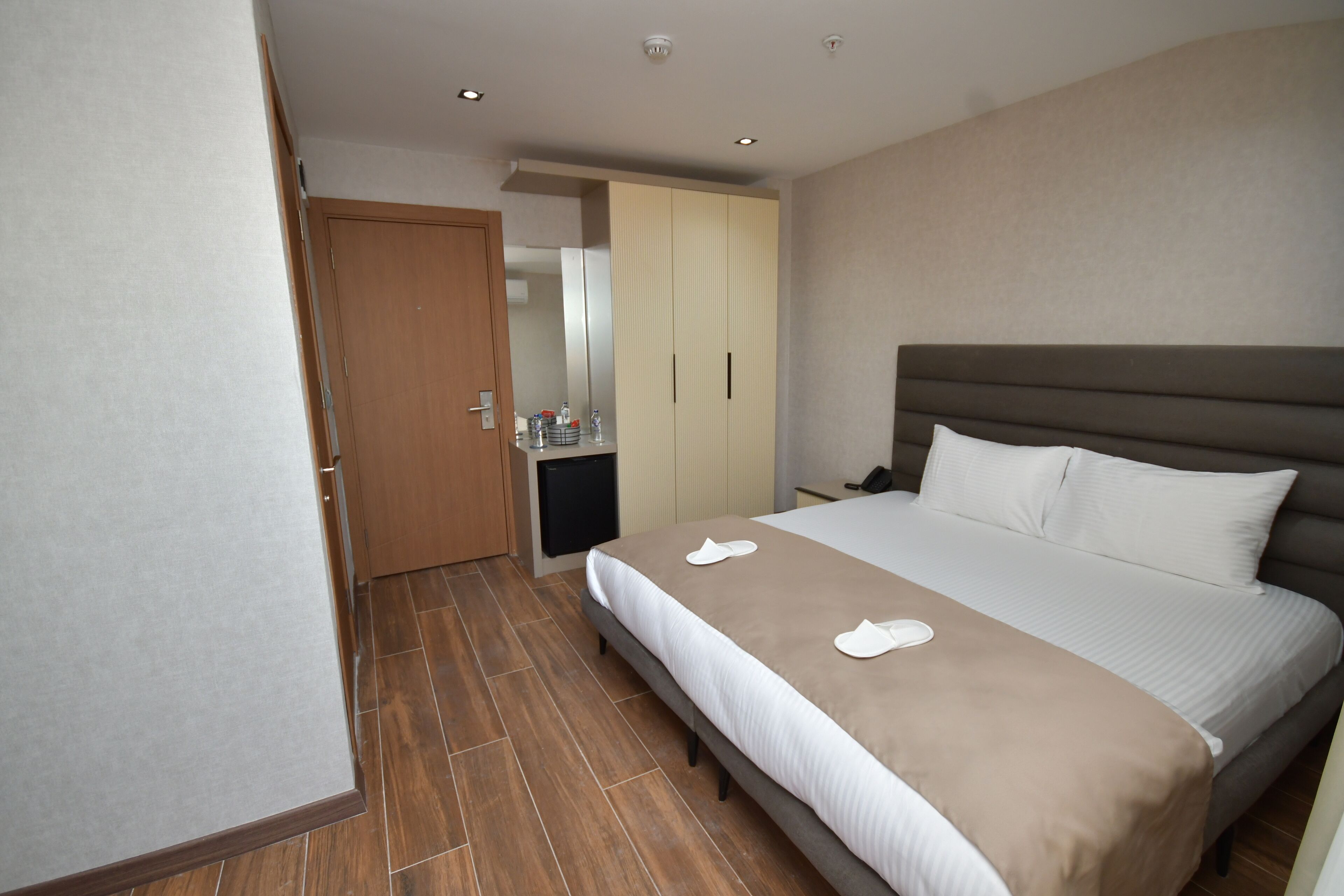A hotel room with a large bed, a wardrobe, a small desk with a chair, and a door leading to another room.