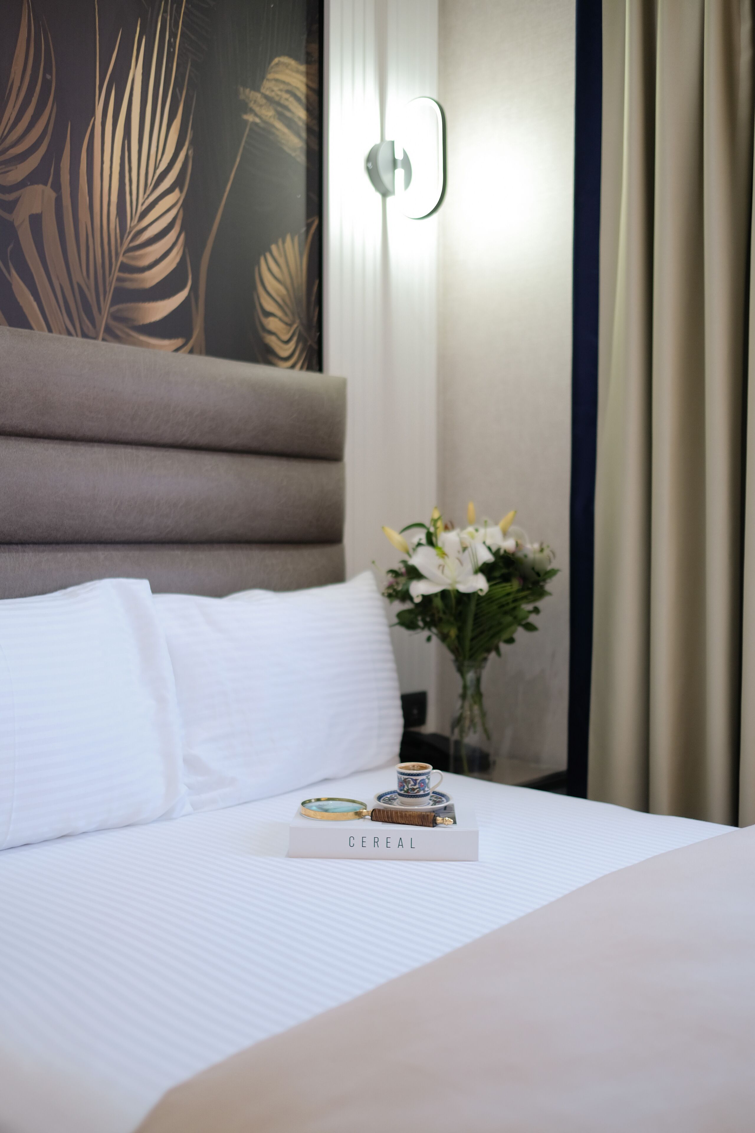 A neatly made bed with white linens, a vase of white flowers, and a tray with a book and a cup.