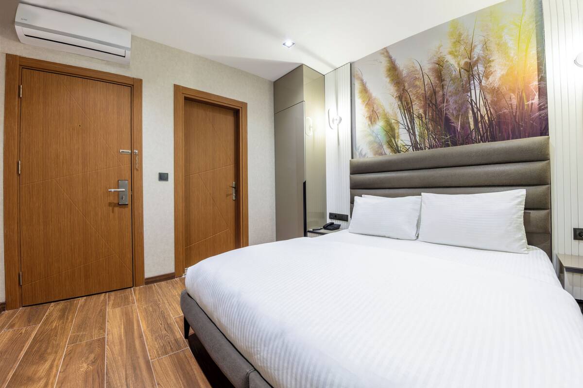 A hotel room with a large bed, two wooden doors, an air conditioner, and a wall-mounted artwork.