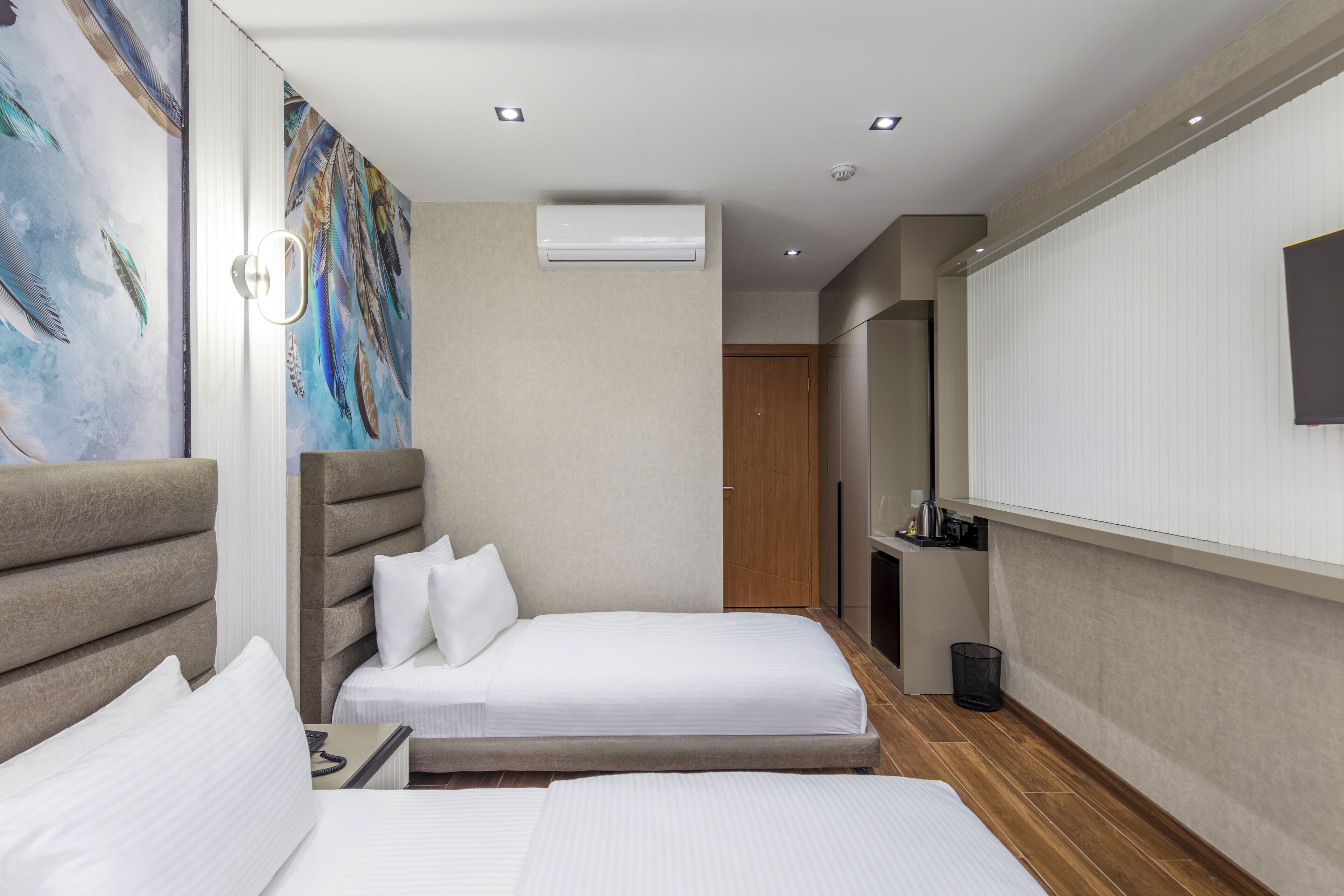 A hotel room with two beds, a painting on the wall, an air conditioning unit, a small desk with a lamp, and a mirror on the wall.