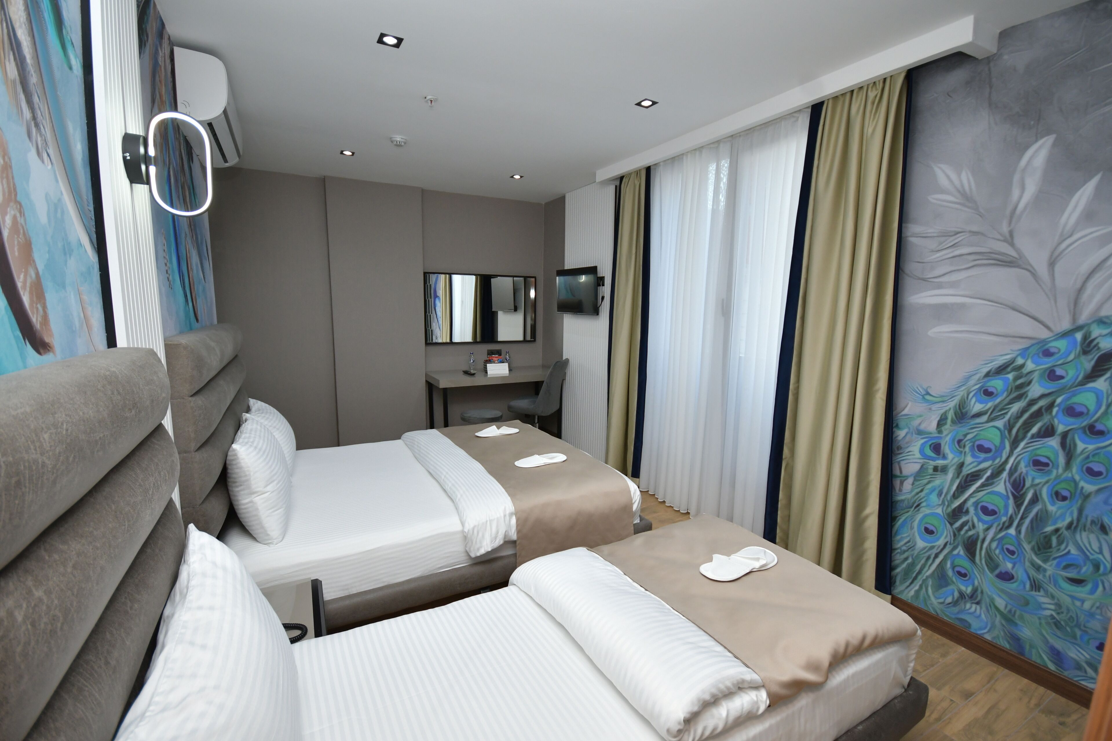 A hotel room with two beds, a desk, a mirror, and a peacock mural.